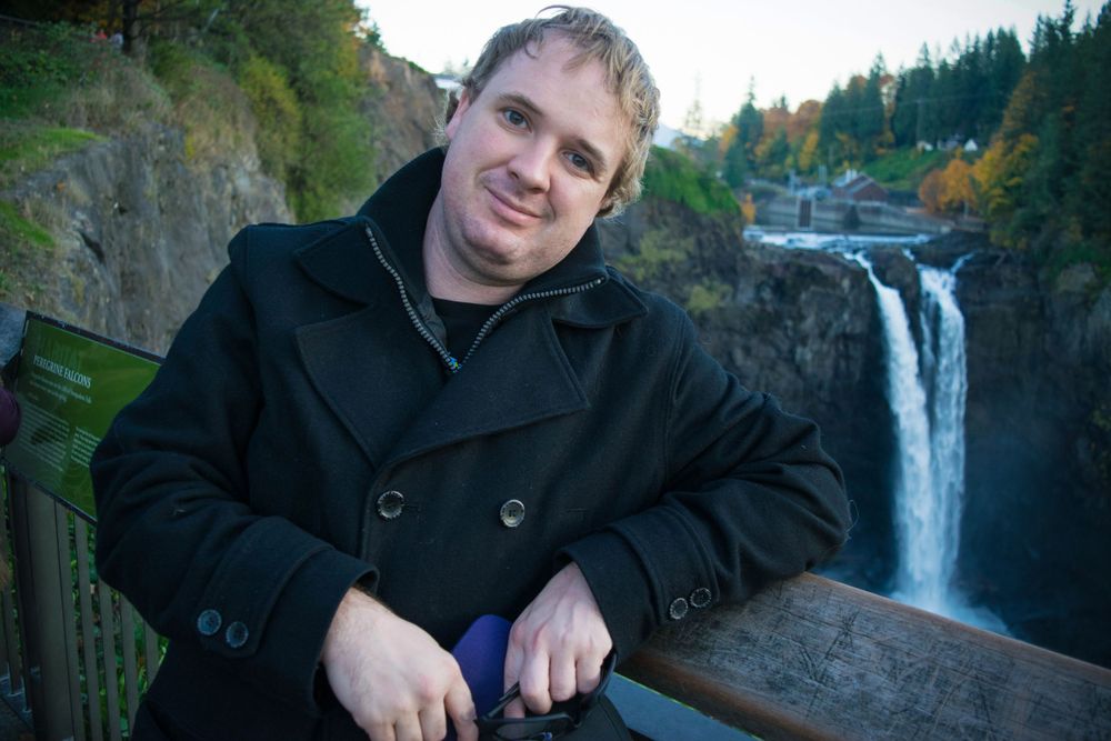 A person in a black coat poses by a scenic waterfall overlook in autumn.