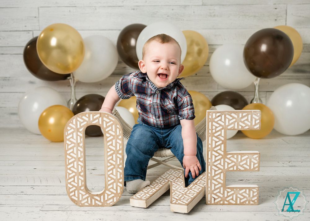 A happy child in plaid sits on decorative 'ONE' letters with gold and brown balloons.