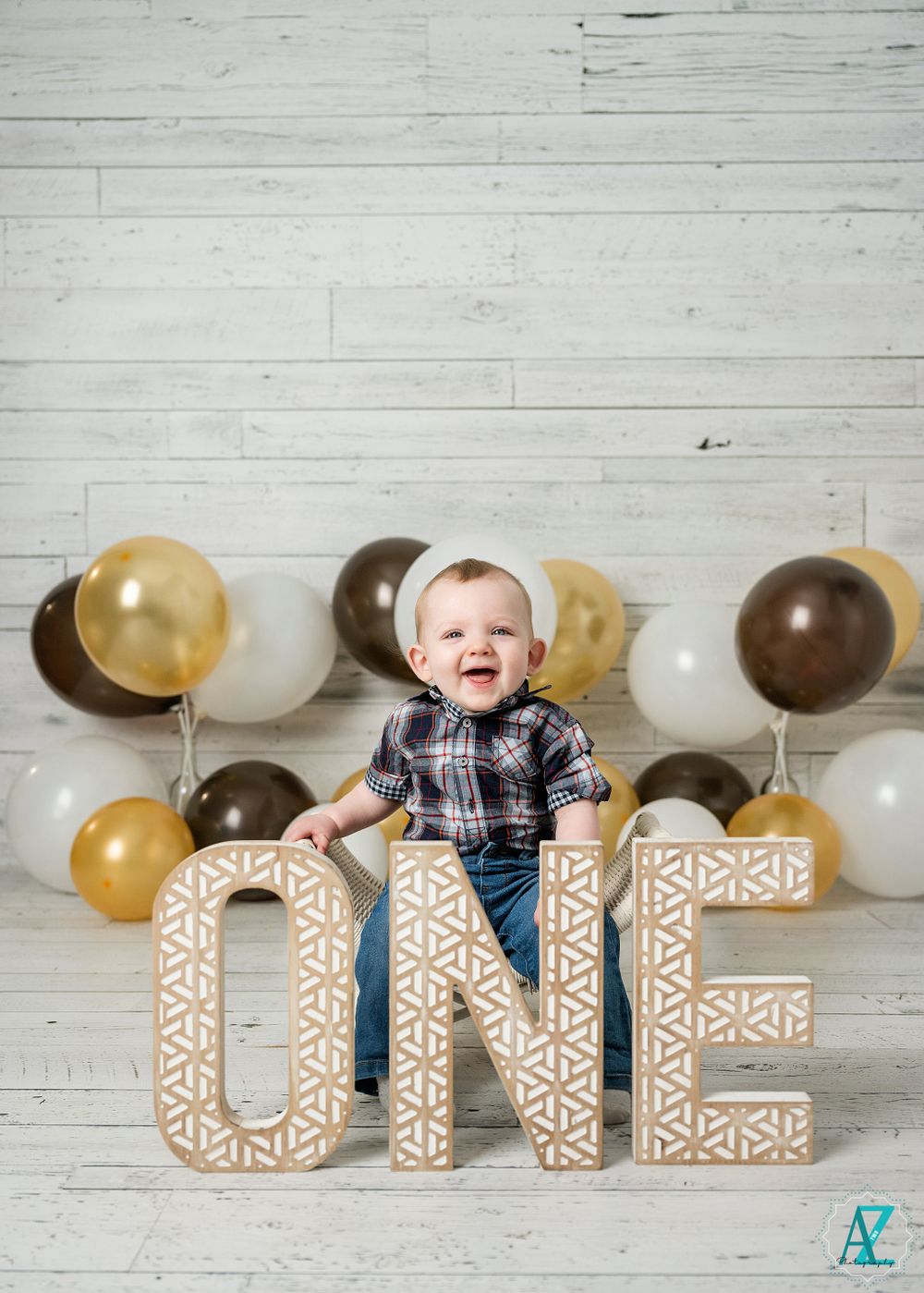 A joyful baby poses with 'ONE' letters and gold and brown balloons against a white wood backdrop.