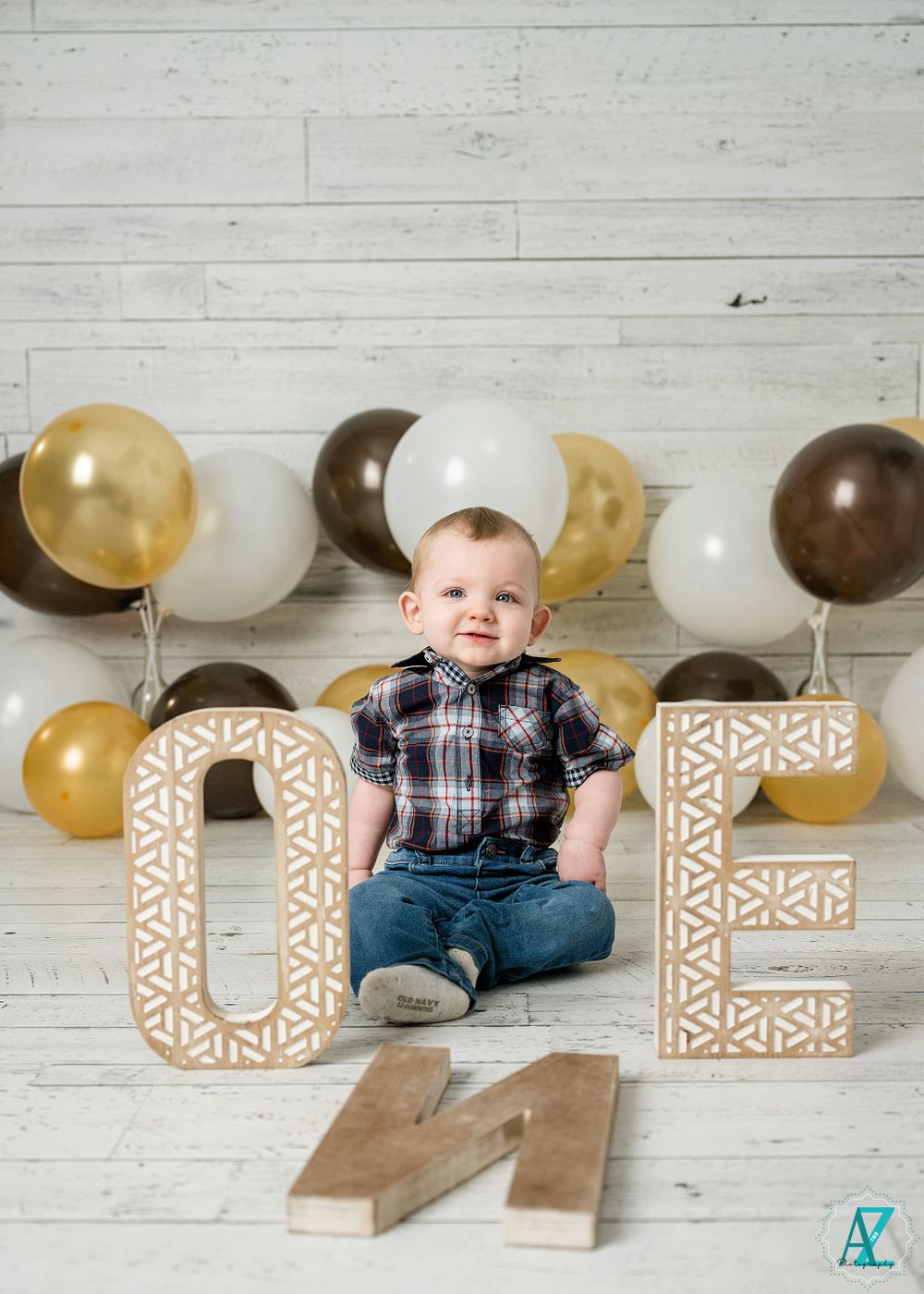 A smiling toddler sits with 'ONE' letters and balloons for a first birthday photoshoot.