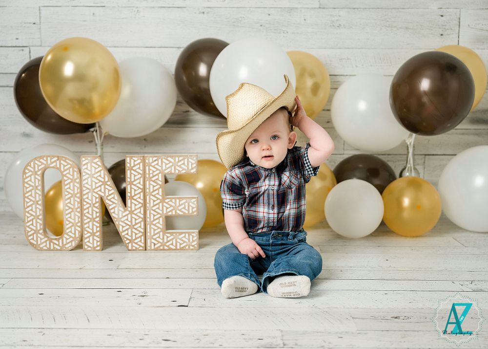 A baby wearing plaid plays with a cowboy hat among gold and brown balloons and 'ONE' letters.