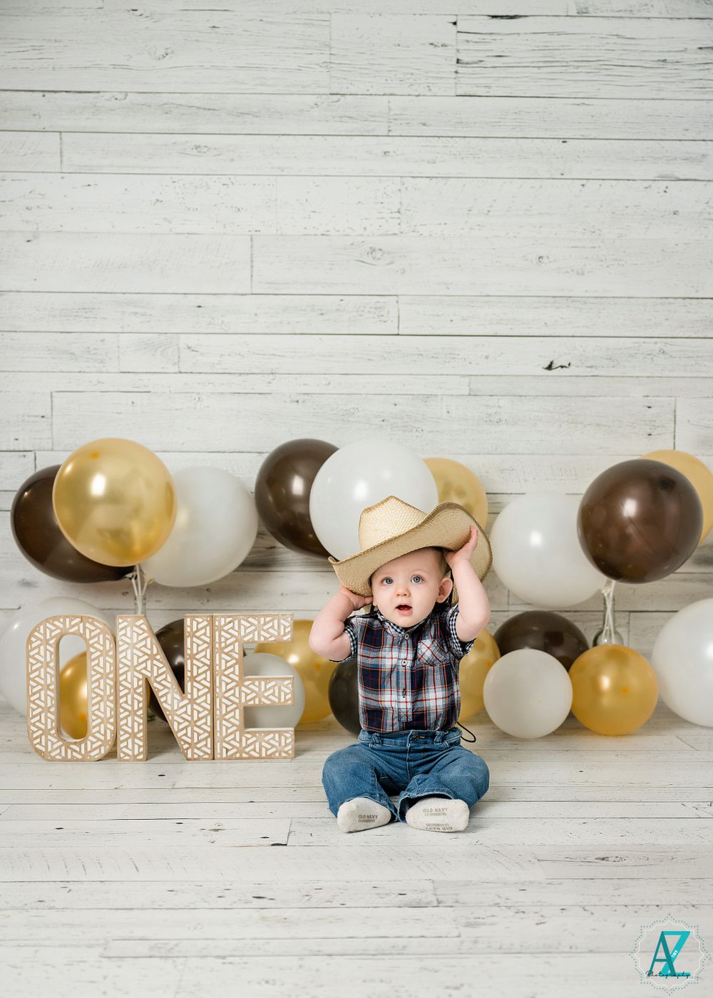 A baby holds a cowboy hat while sitting by 'ONE' letters and festive balloons.