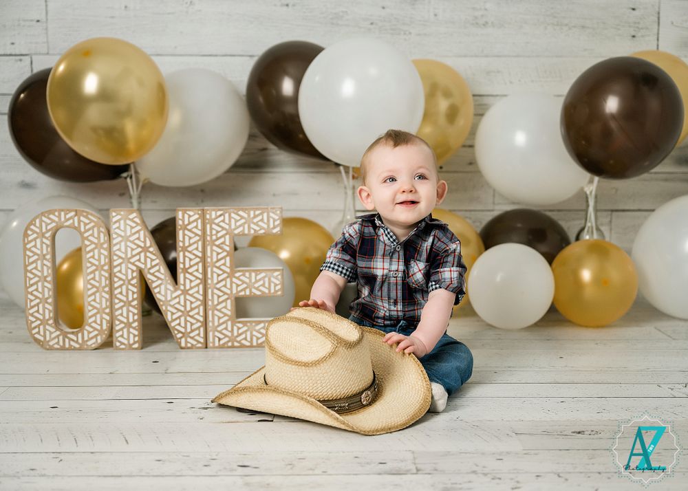A baby in plaid sits with a cowboy hat surrounded by celebratory balloons and 'ONE' decor.