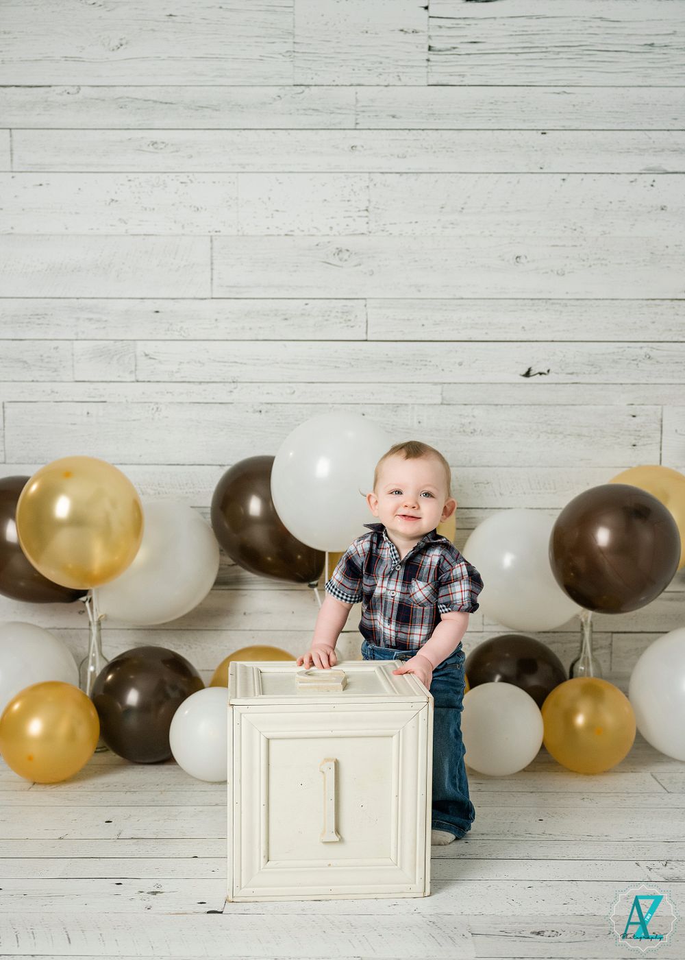 A baby stands by a white cube with gold and brown balloons in a rustic setting.
