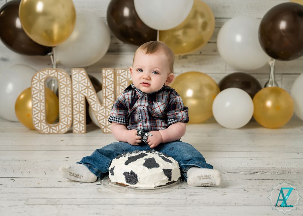 First birthday setup featuring decorative ONE letters, balloons, and cow print smash cake on white wooden floor.