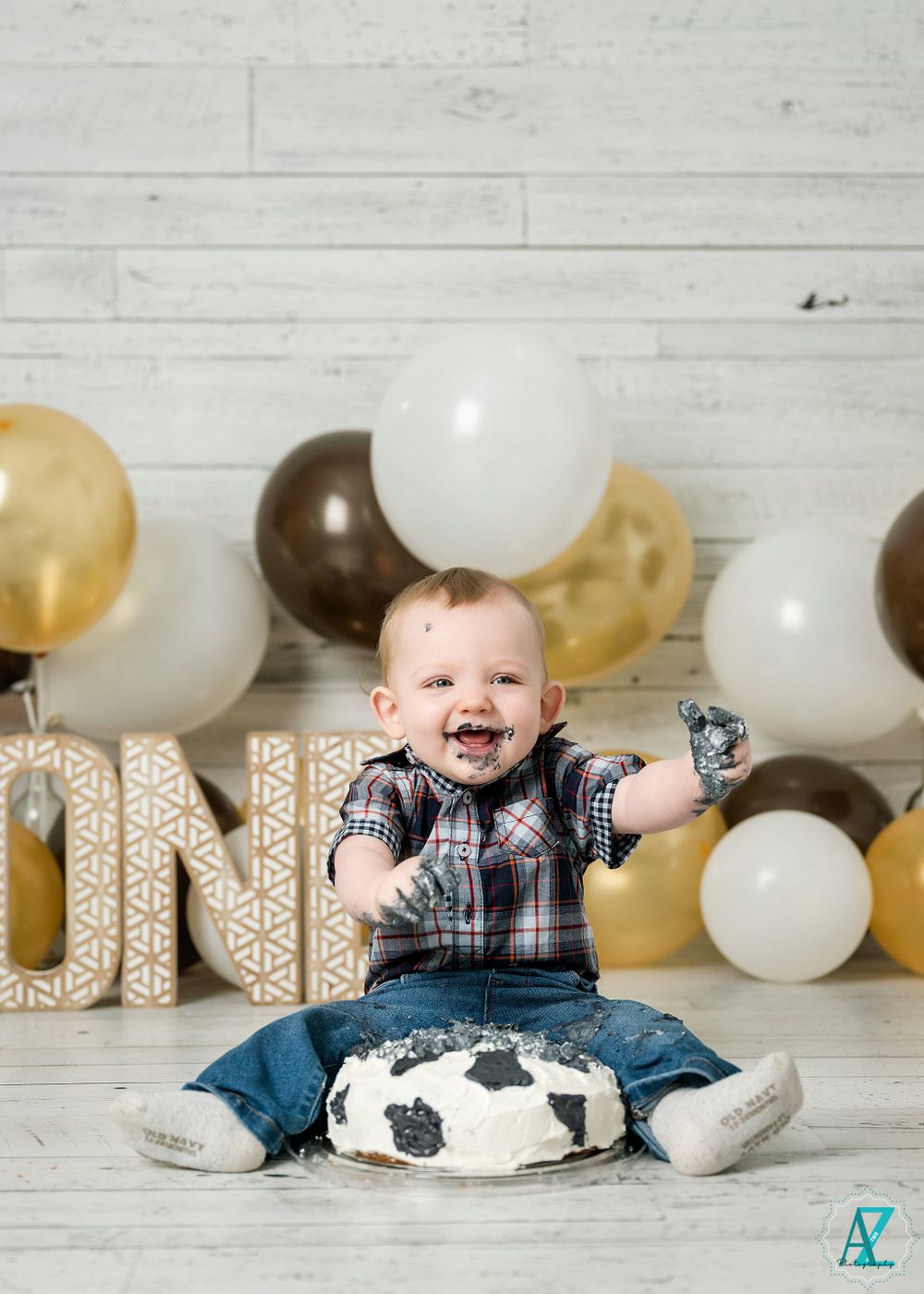 First birthday celebration with gold letter decorations and brown and white balloons against shiplap background.