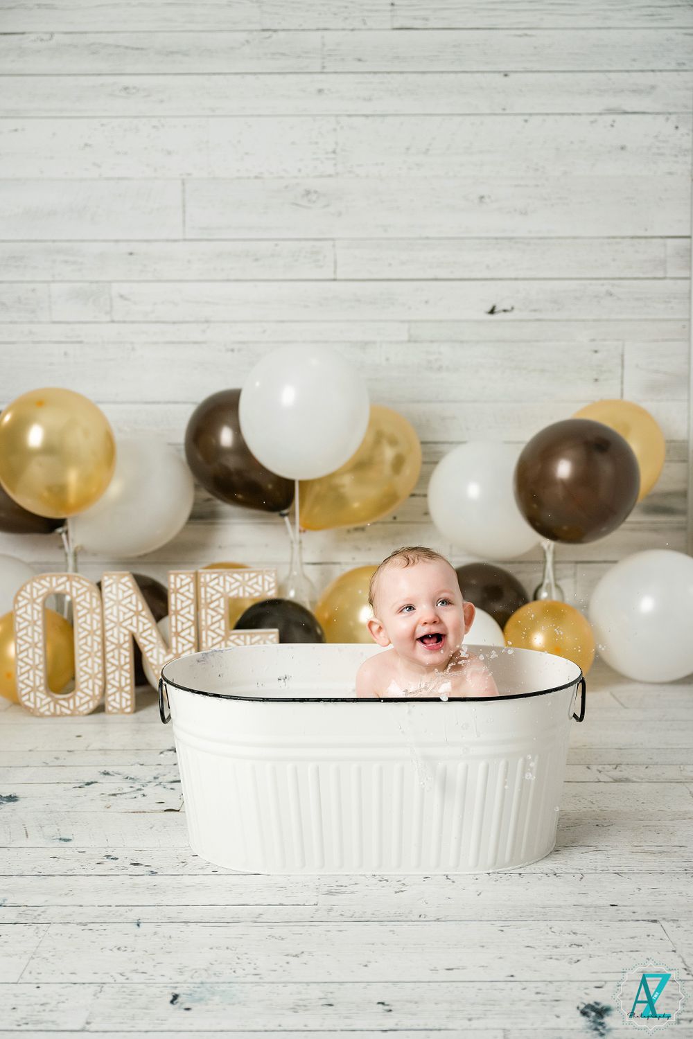 A baby celebration setup with gold and white balloons and a white basin against a rustic wooden wall background.