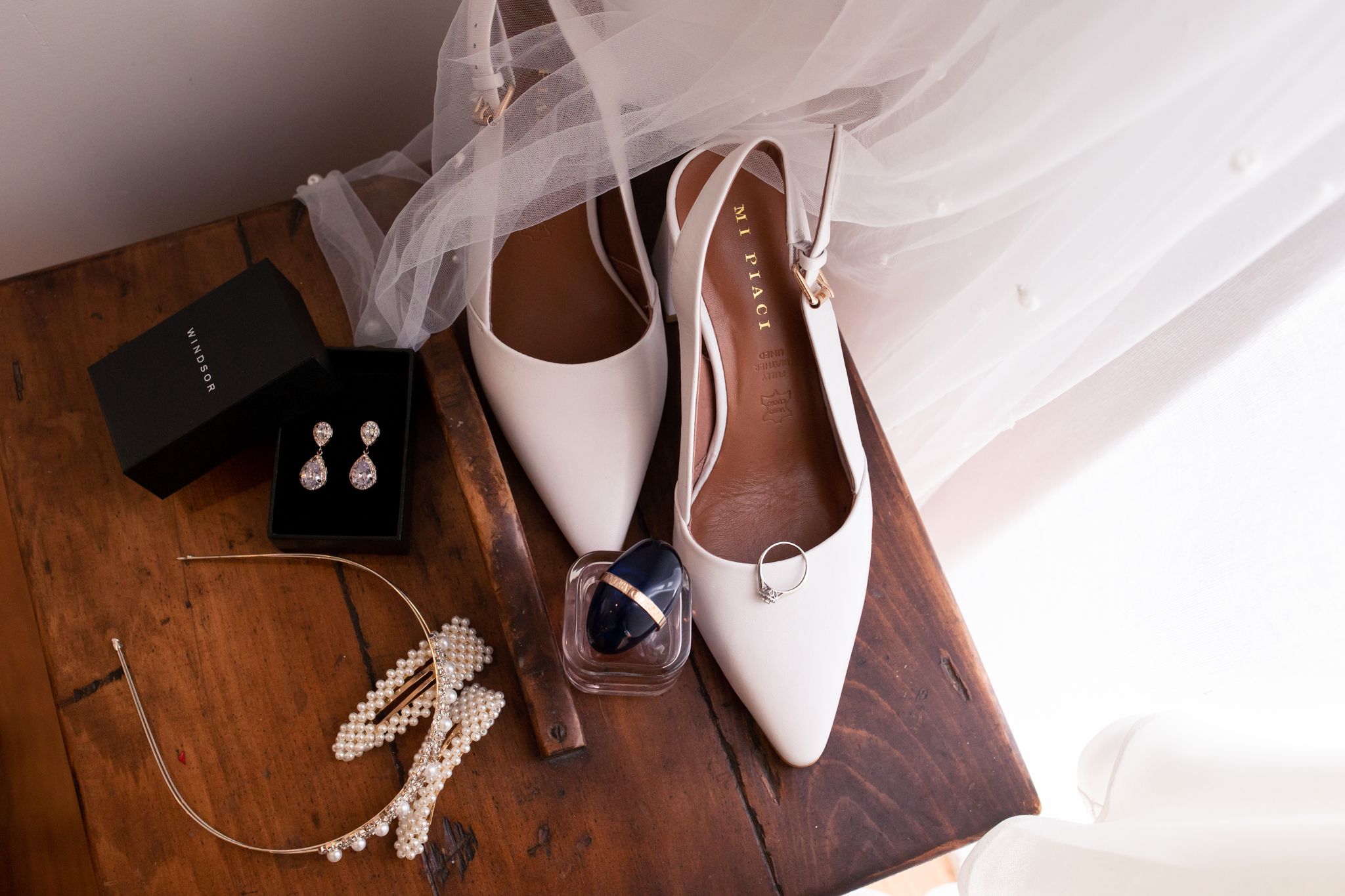 Wedding accessories including white pointed shoes, jewelry and a ring box on wooden surface.