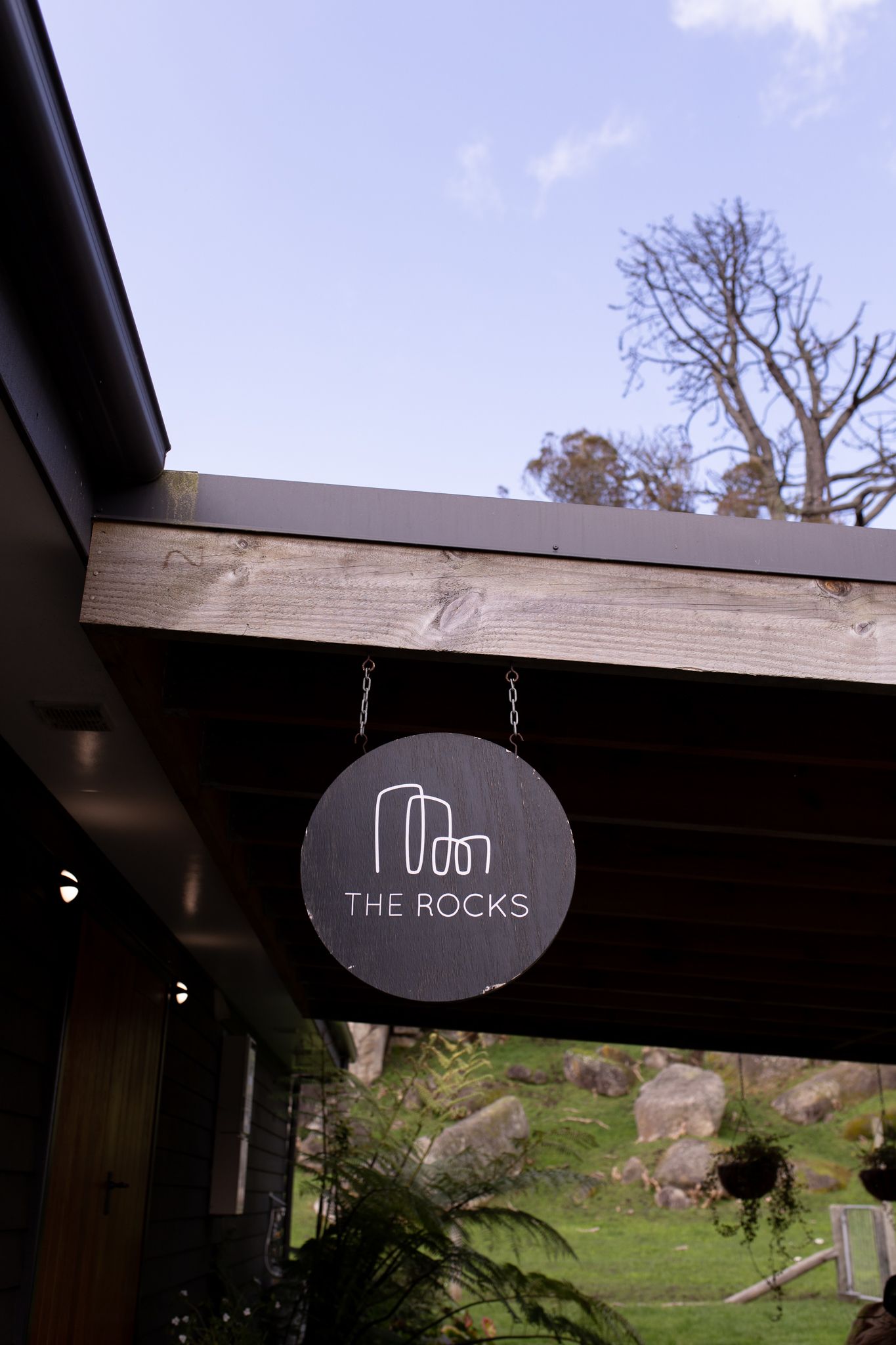 The Rocks venue sign hanging from wooden beam against blue sky.