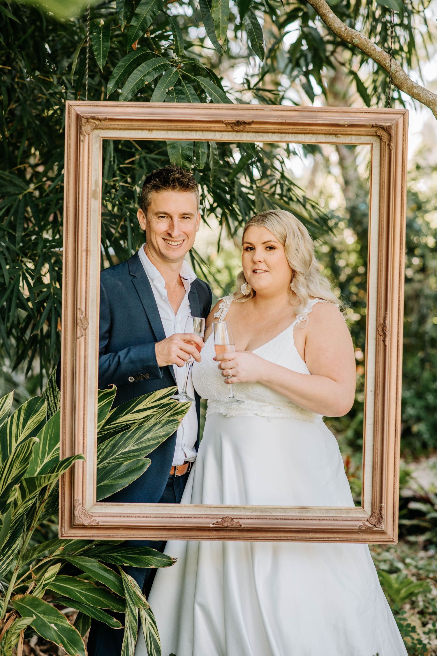 Wedding couple poses inside gold picture frame surrounded by lush tropical greenery while holding champagne flutes.