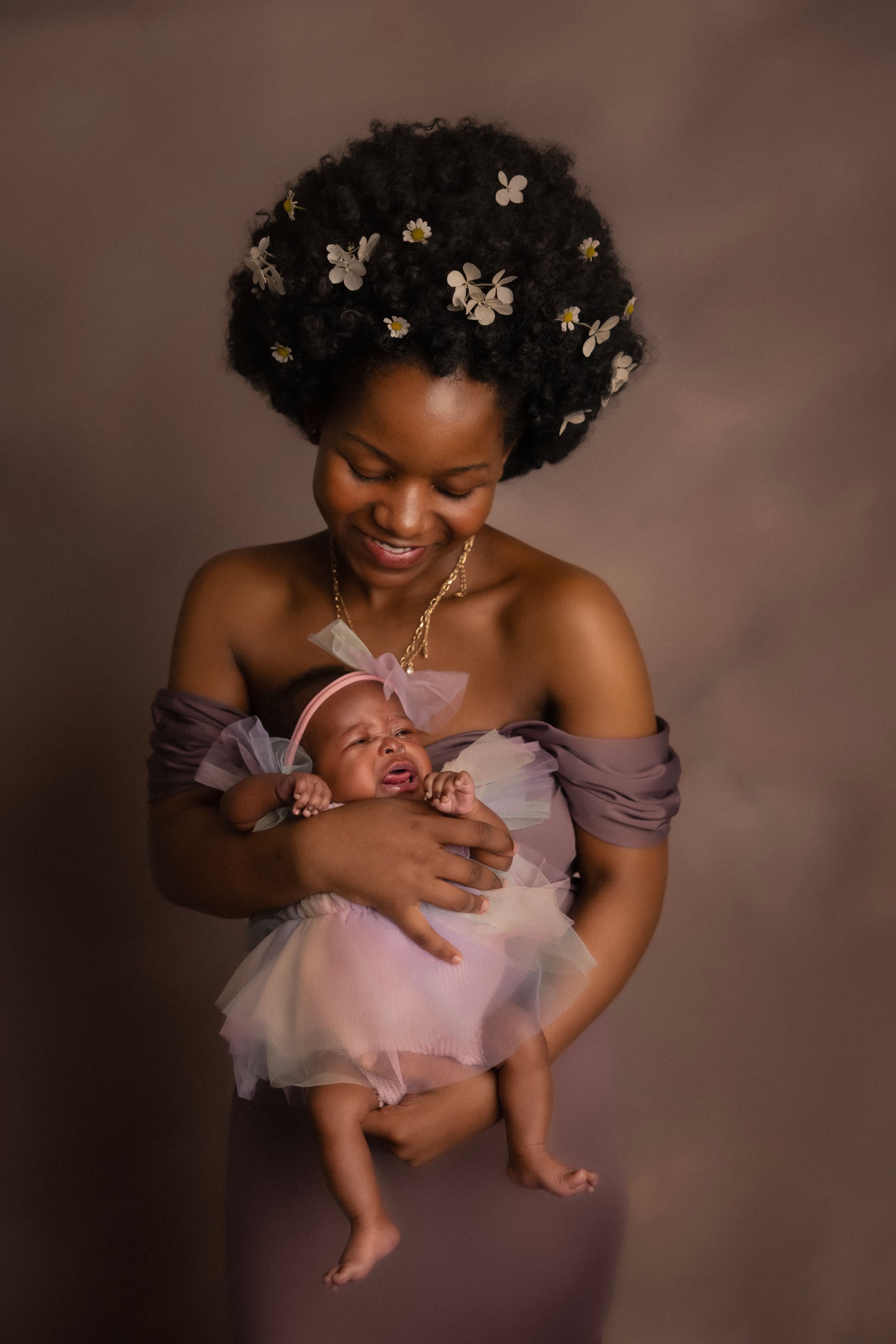 Smiling Black mother with floral afro holds her newborn baby dressed in a pink tutu for a studio portrait.