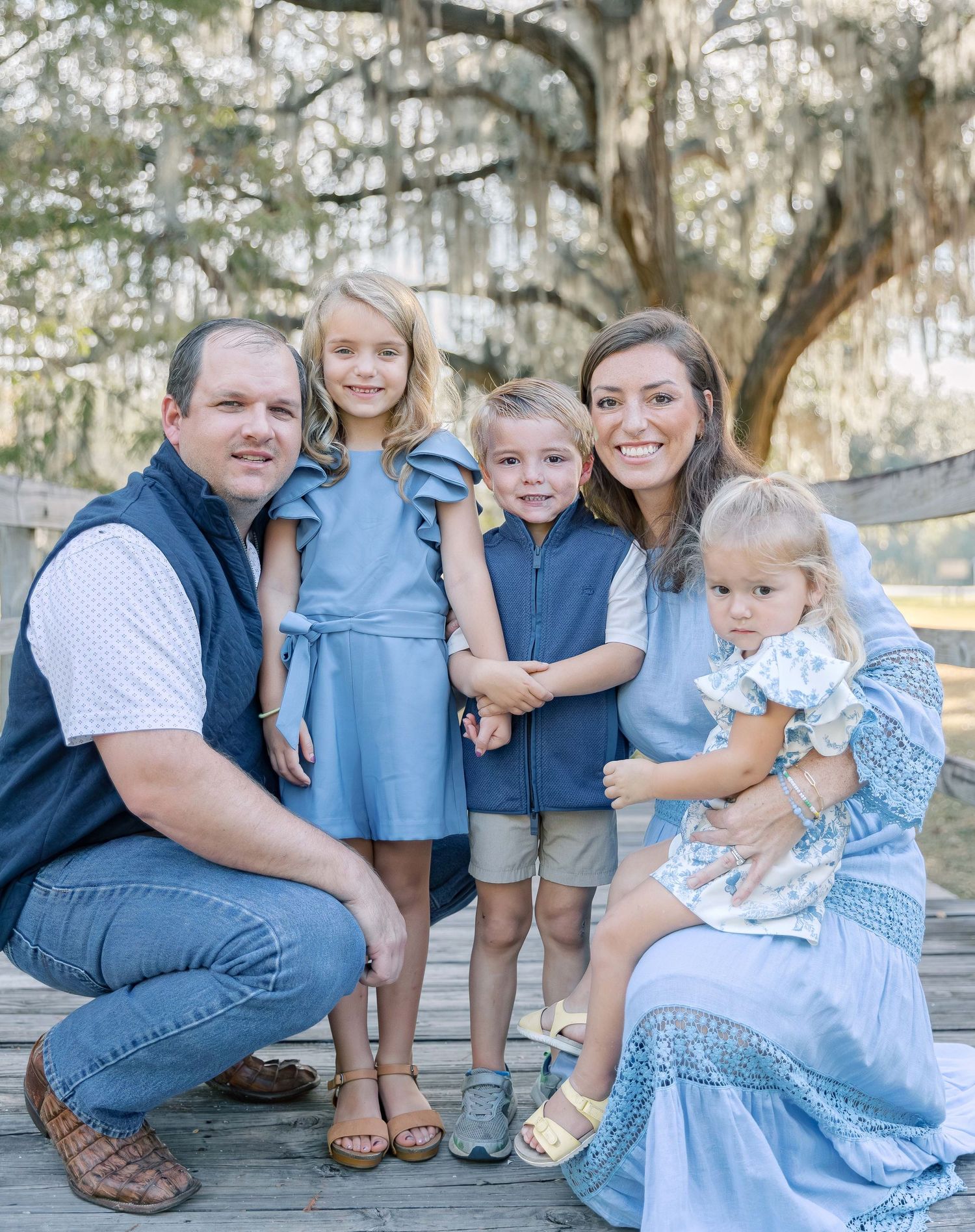 A group poses in coordinated blue outfits on wooden steps under a Spanish moss draped oak tree.