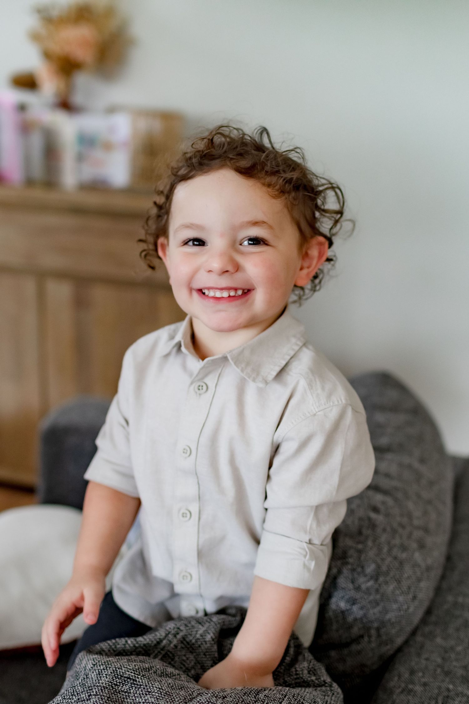 A joyful child in a light colored button-up shirt sits on a grey couch.