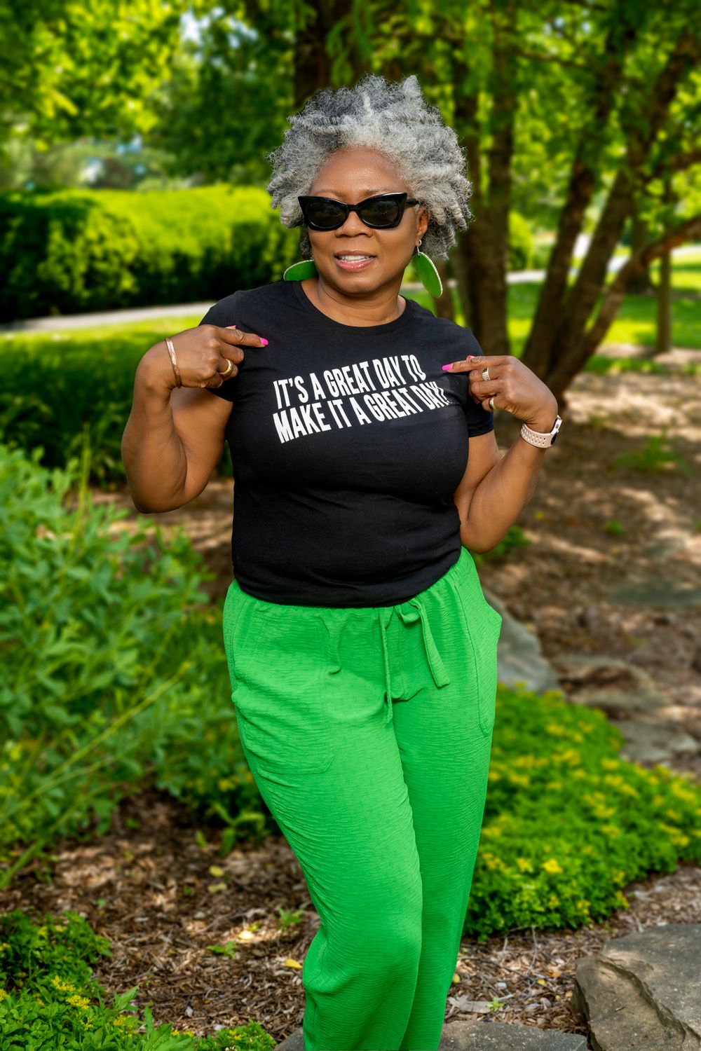 Woman in black t-shirt and bright green pants poses cheerfully in sunny park setting.