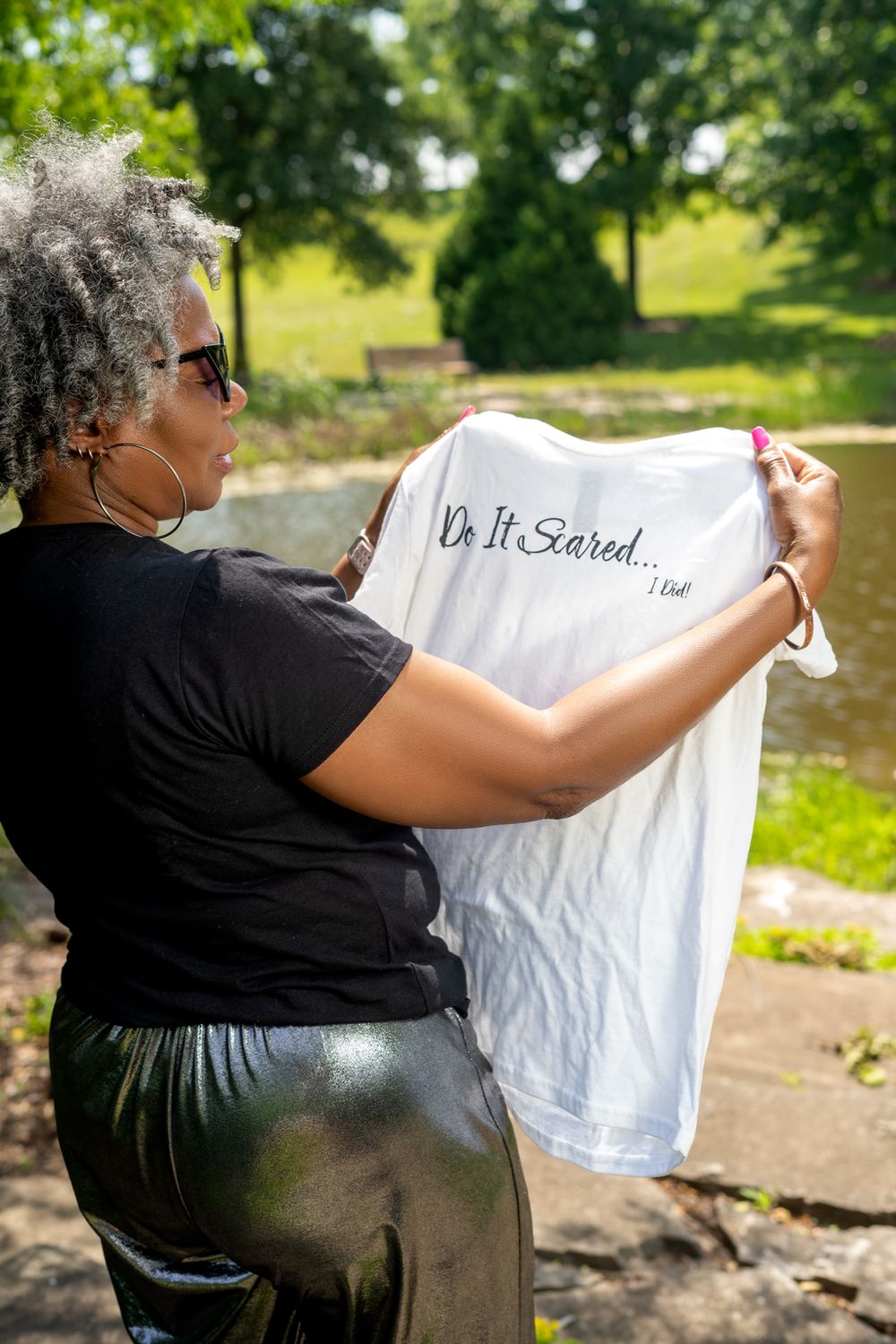 Person in sunglasses holds up a white t-shirt with text in an outdoor garden setting.