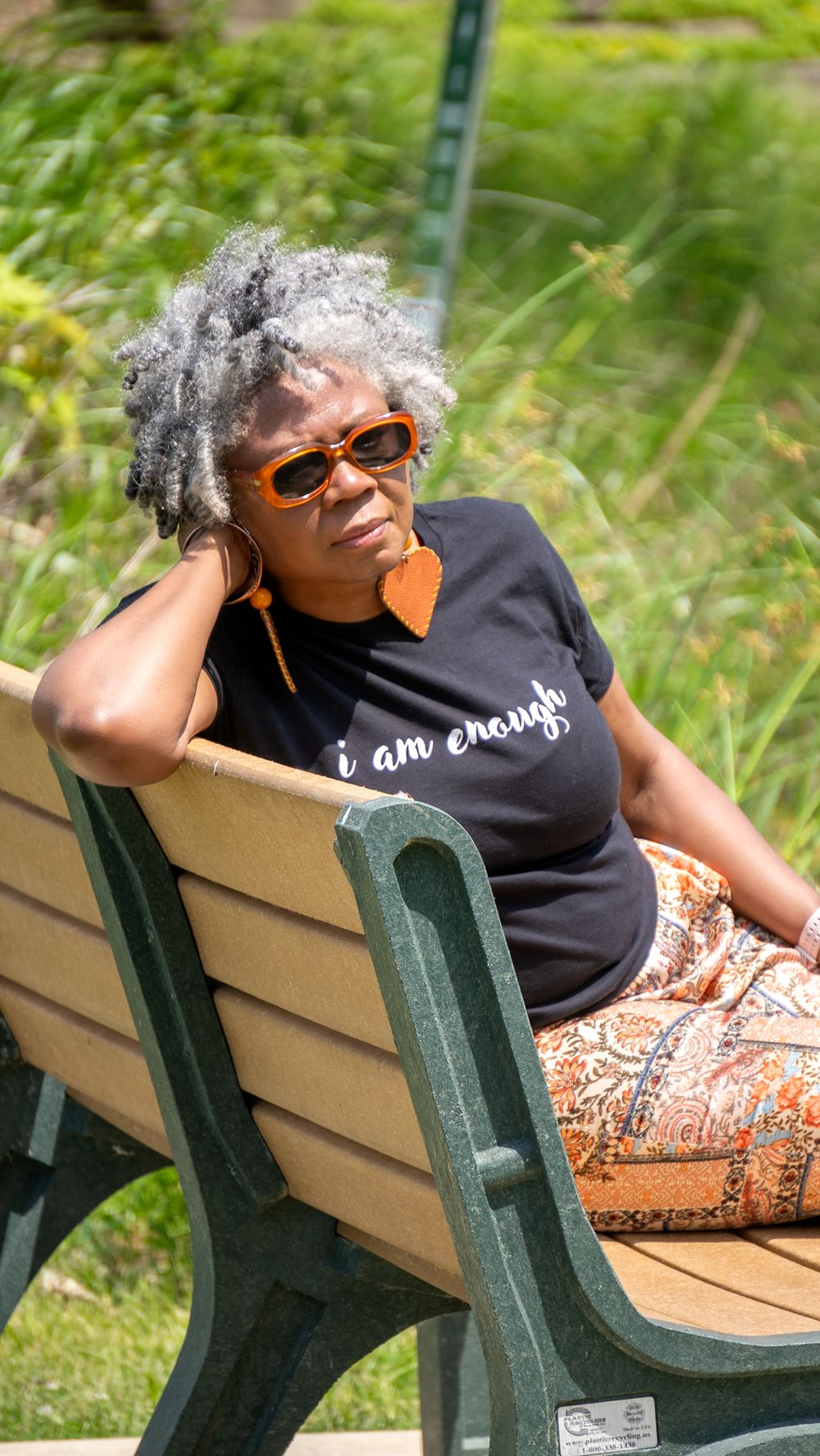 Person in black t-shirt and orange earrings relaxes on a park bench on a sunny day.