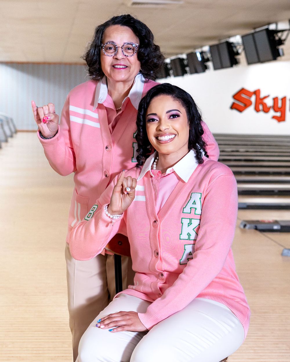 Two people wearing matching pink letterman jackets pose together at a bowling alley.