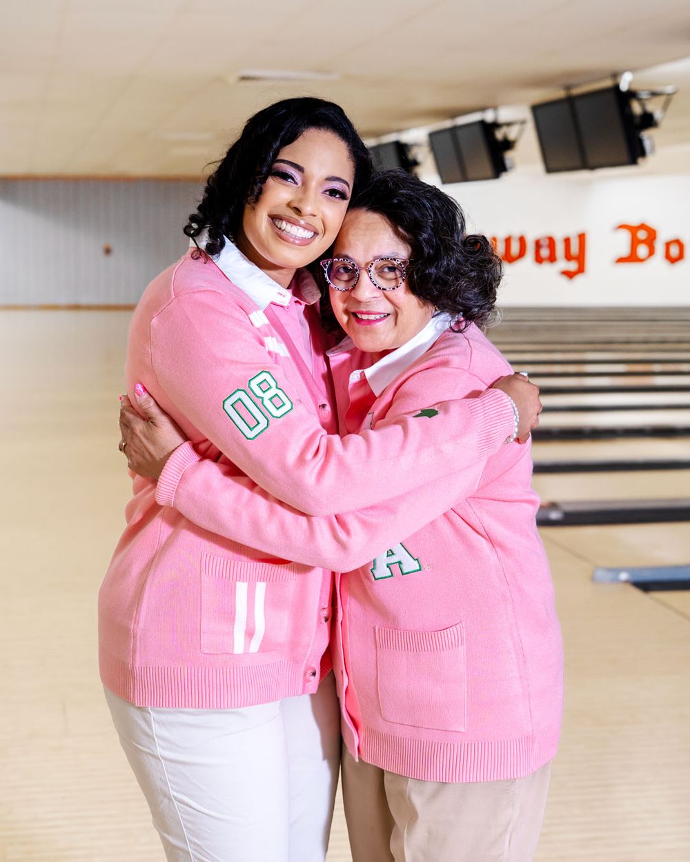Friends in matching pink varsity jackets share a warm embrace at a bowling alley.
