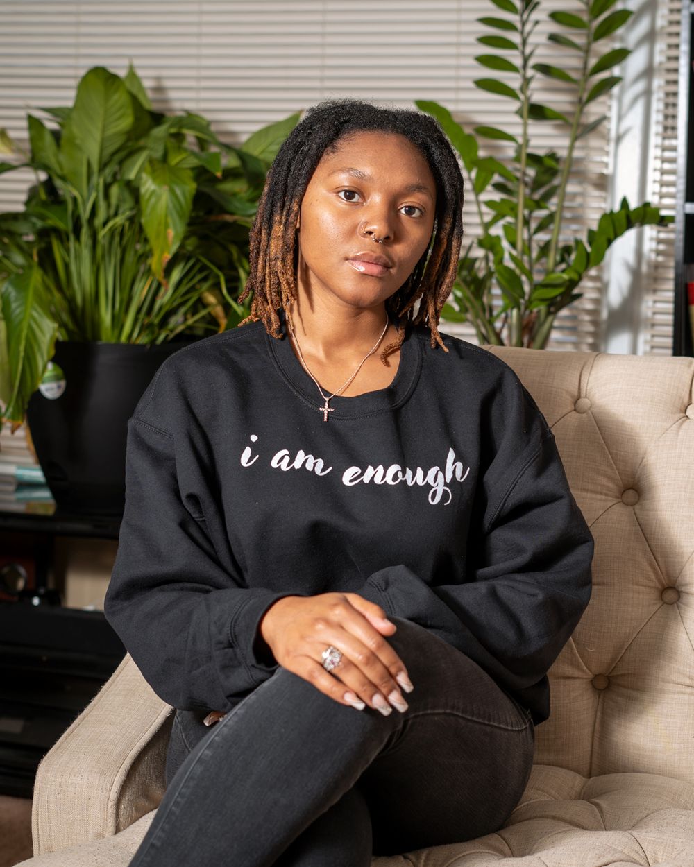 Relaxed pose on beige couch wearing black 'I am enough' sweatshirt with plants in background.