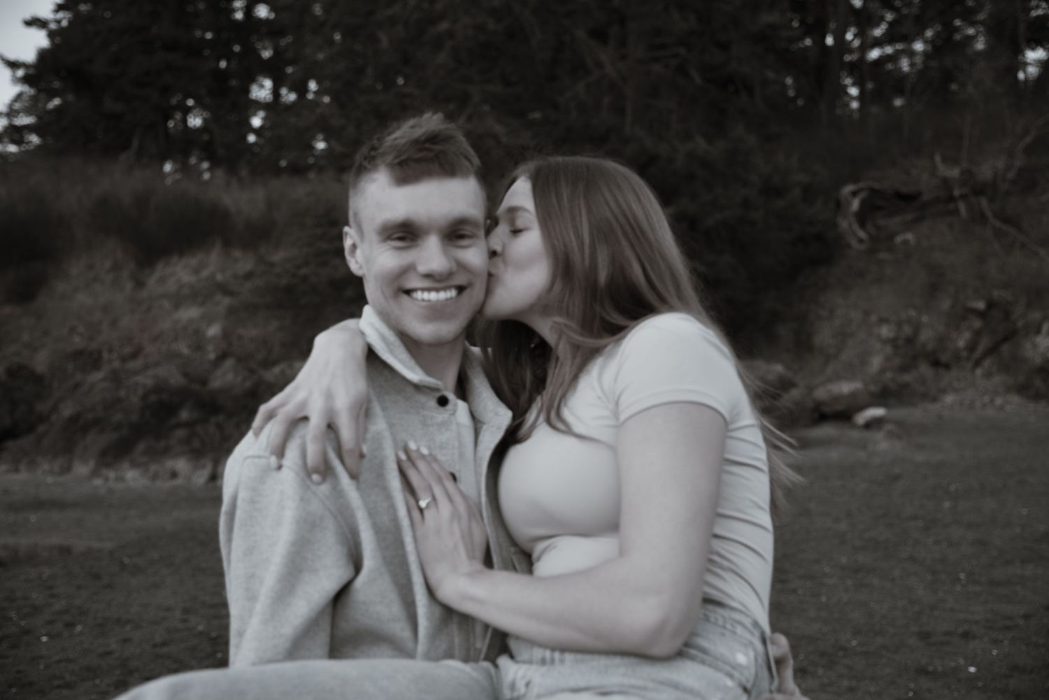 Black and white photo of smiling man receiving a kiss on the cheek from a woman outdoors near trees.