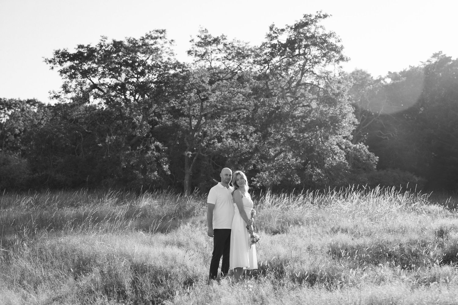 Black and white photo of smiling man receiving a kiss on the cheek from a woman outdoors near trees.