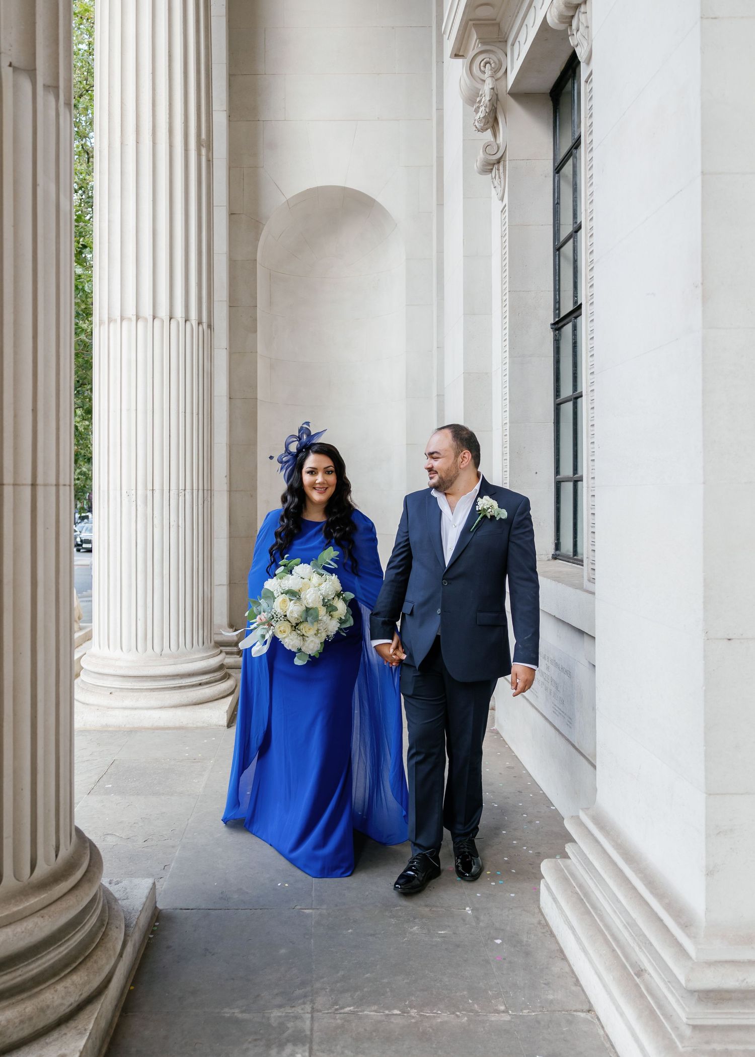 A couple poses between classical columns, with one wearing a royal blue gown holding a floral bouquet at a downhill wedding in London.
