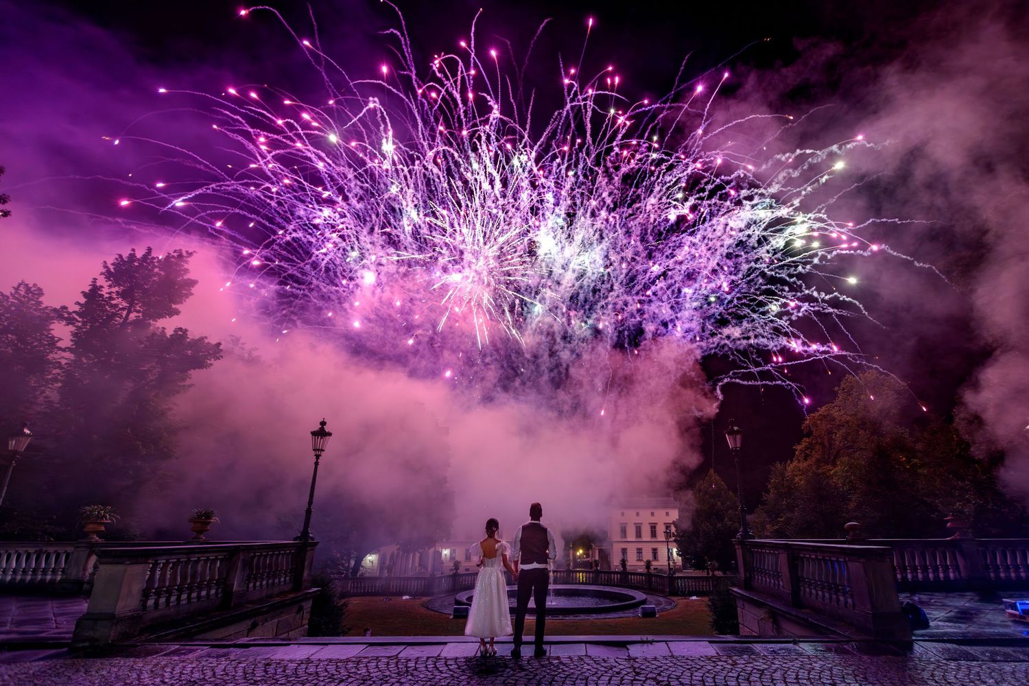 Pink and purple fireworks illuminate the night sky over a city with buildings silhouetted against the bursting display.