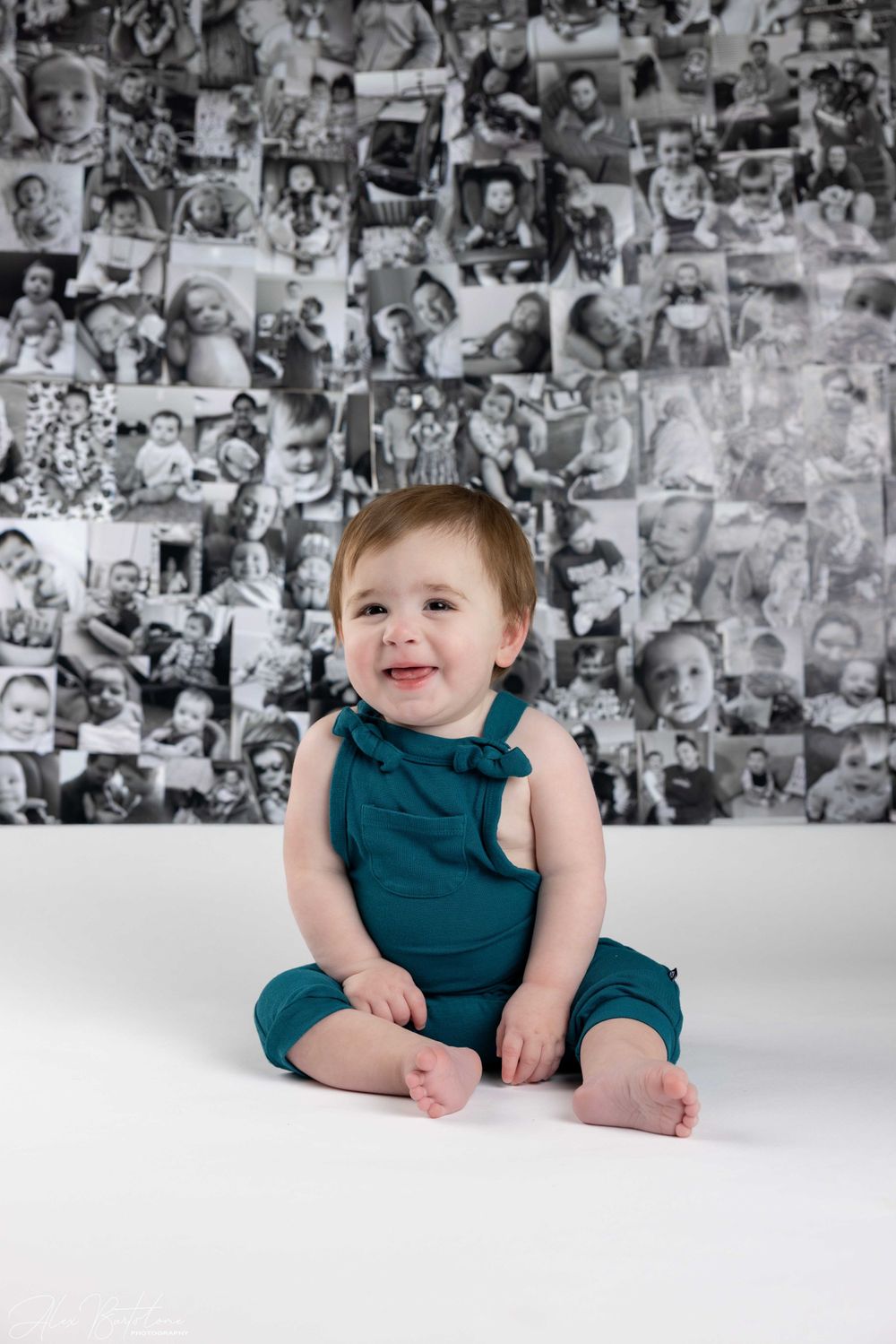 Happy baby in teal romper sits on white surface before black and white photo wall.