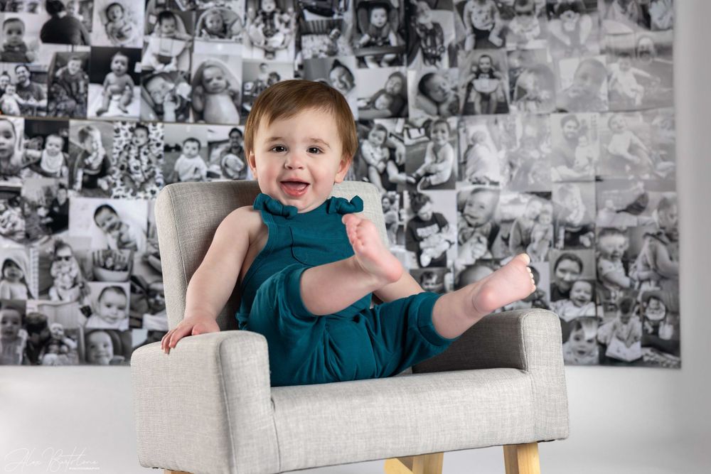 Laughing baby in teal dress sits in gray chair before black and white photo collage wall.
