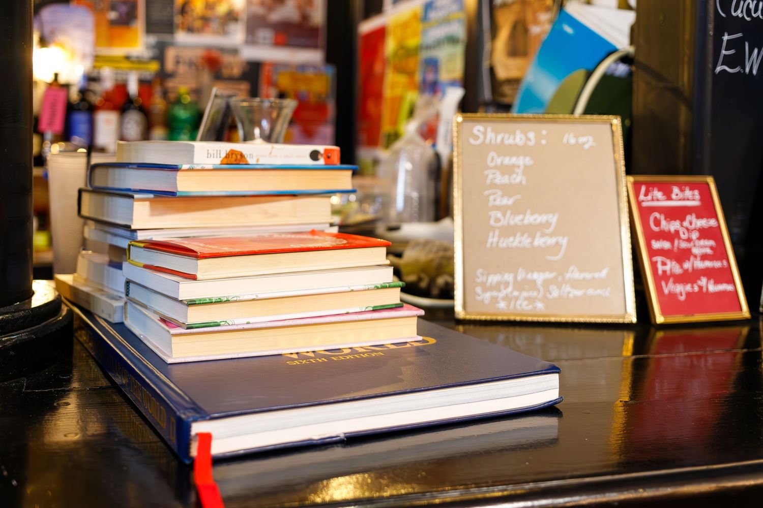 Stack of books arranged on a dark wooden counter top in a cafe setting.