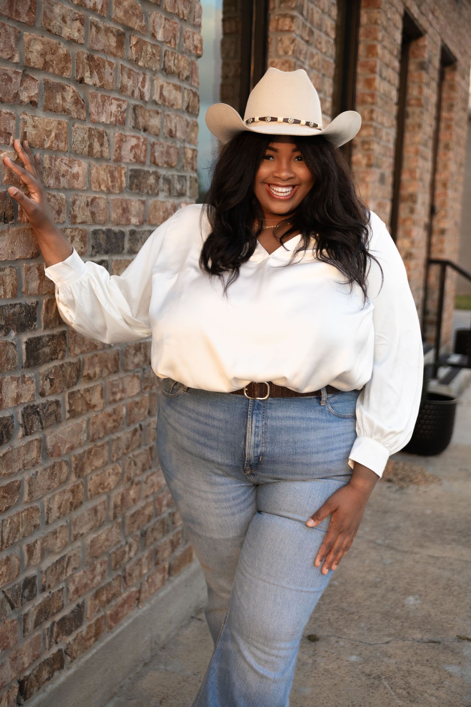 Model in white puffy sleeve top and jeans poses playfully against brick wall.