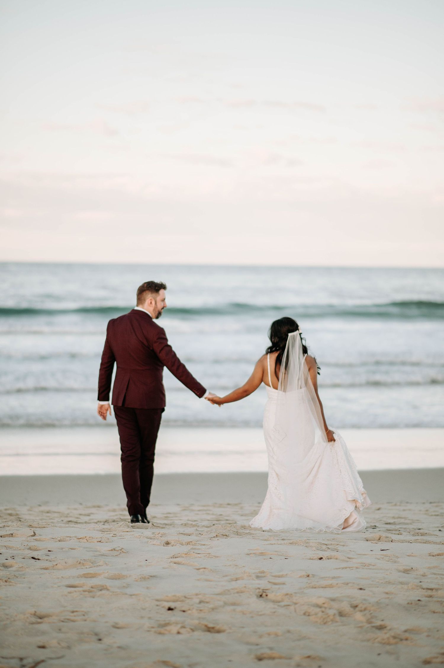 Newlyweds walking hand in hand on beach at sunset in formal attire.