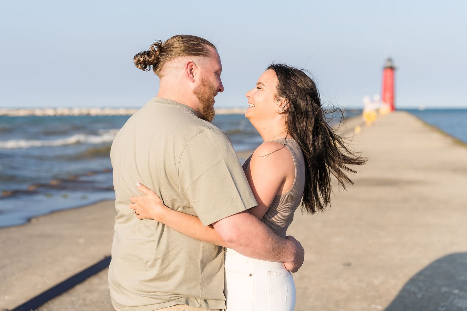 Romantic couple embracing on a pier with a red lighthouse and blue ocean in the background on a sunny day.