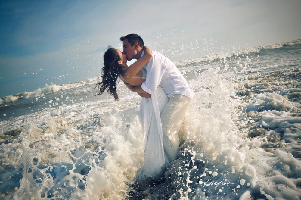 Romantic couple embracing in white attire as ocean waves splash around them on the beach.