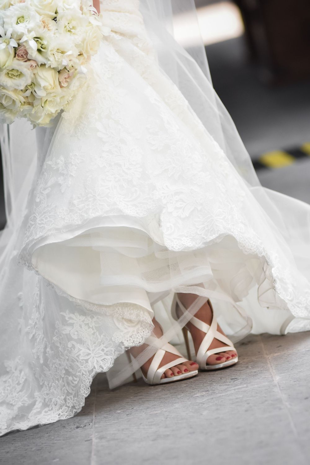 Close-up of white lace wedding dress hem and strappy bridal sandals on grey floor.
