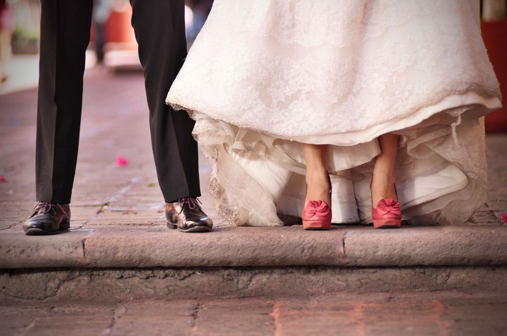 Close-up of bride in red shoes and groom's feet on stone pavement.