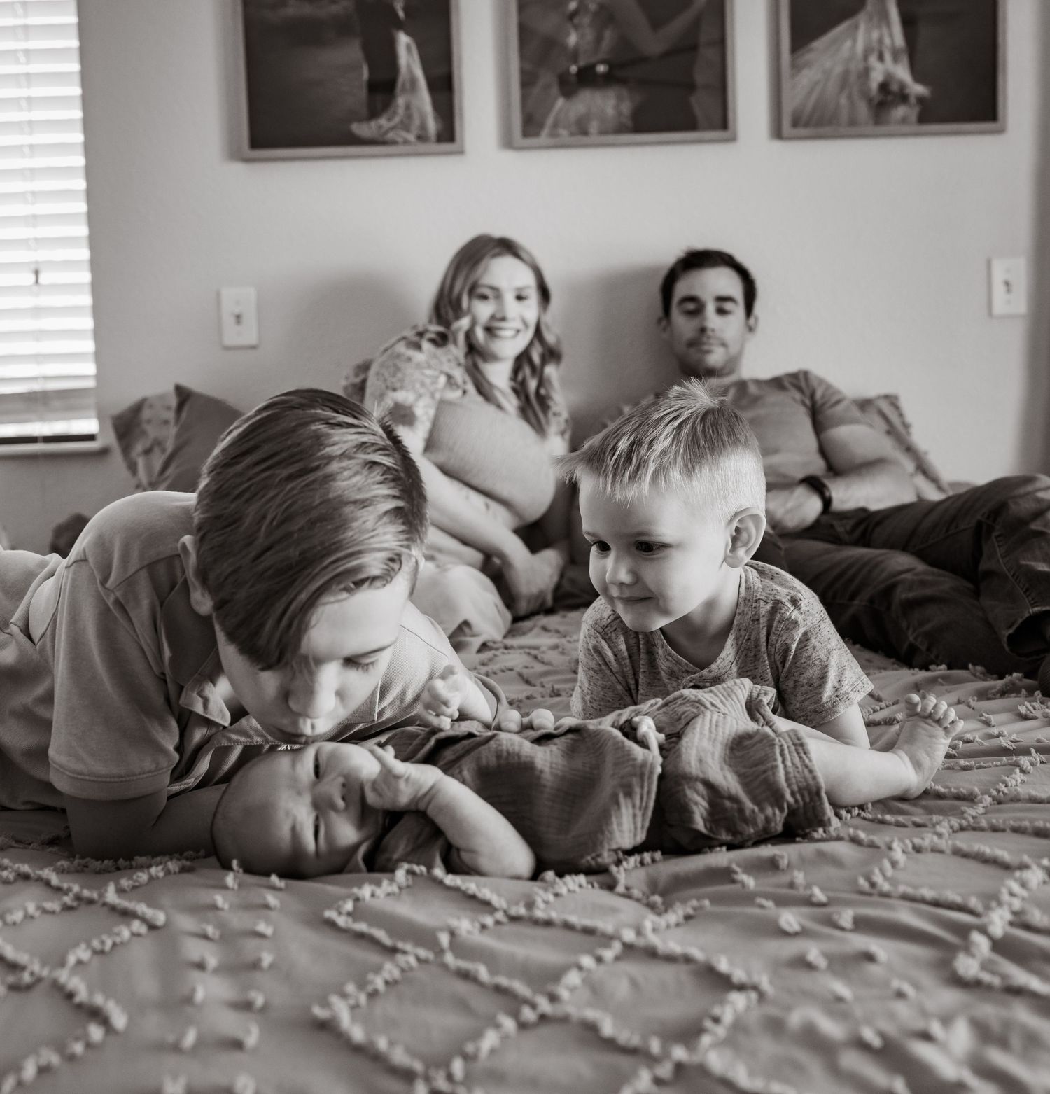 A black and white family photo shows a cozy moment on a bed with framed photographs visible on the wall behind them.