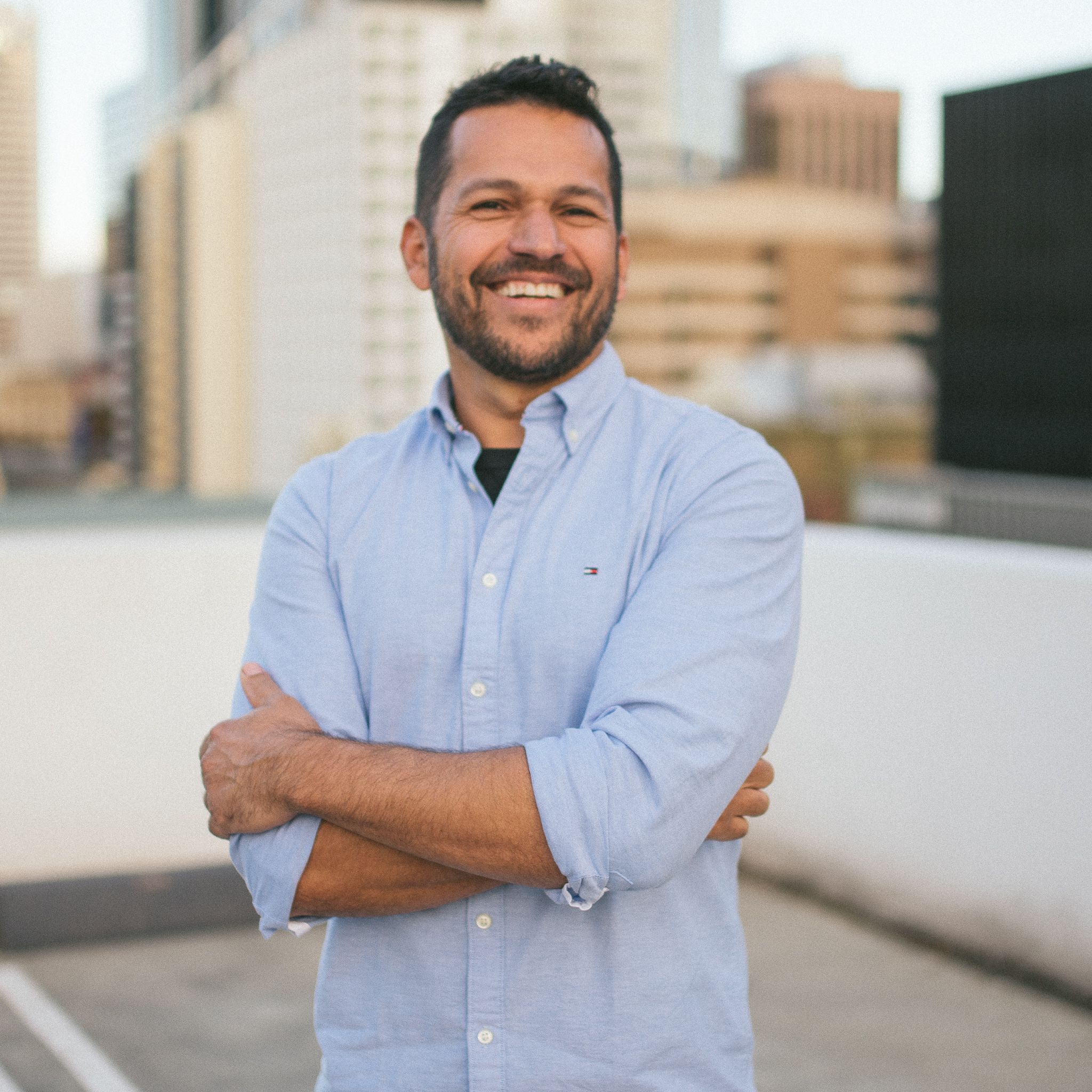 Professional headshot outdoors with city buildings in background.