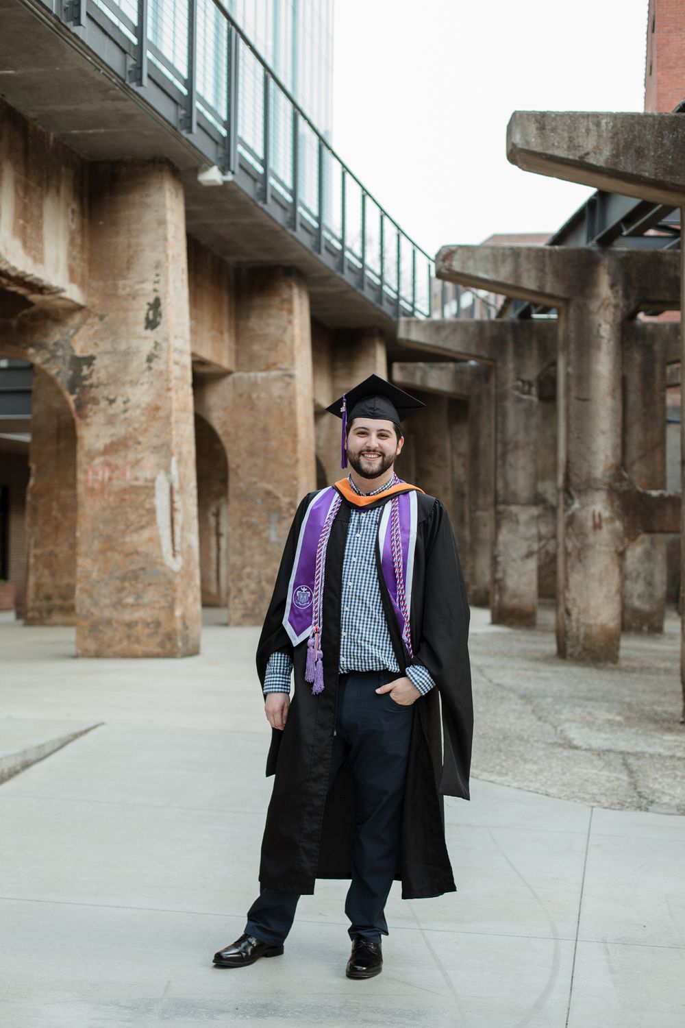 Graduate in cap and gown with purple stole poses in front of architectural columns.