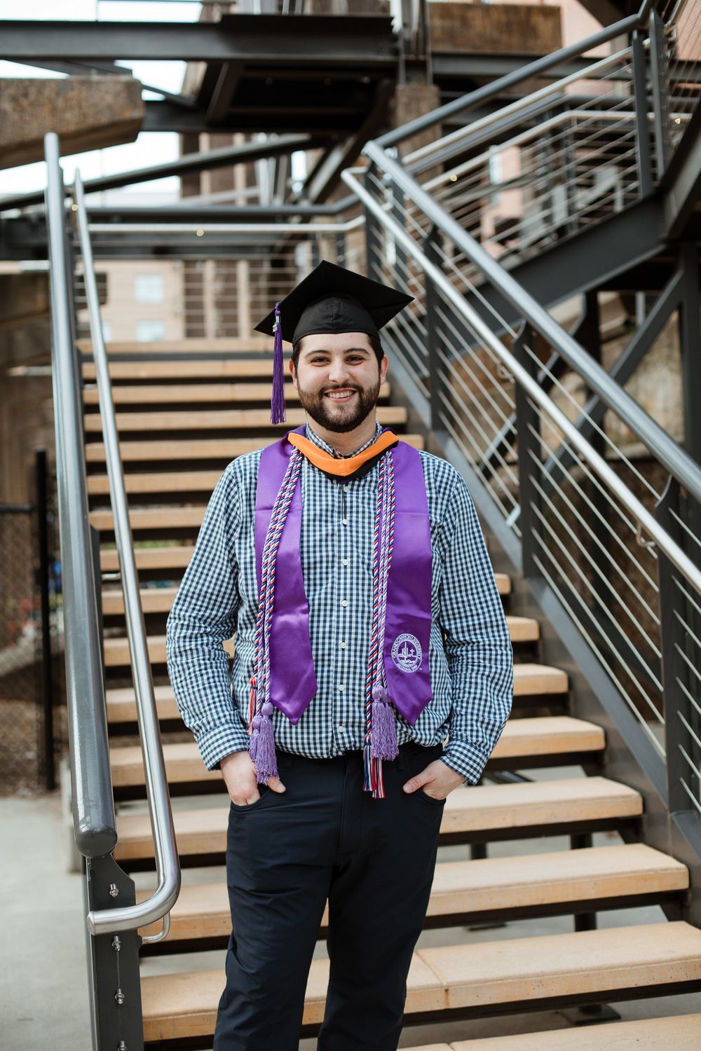Graduate in checkered shirt and purple stole poses on modern wooden staircase.
