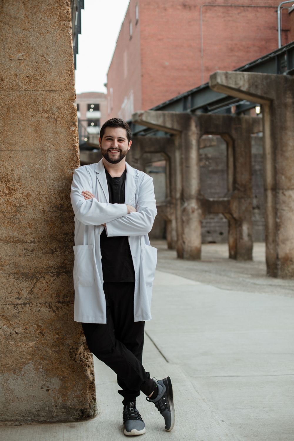 Professional wearing white coat poses against textured stone columns in urban setting.