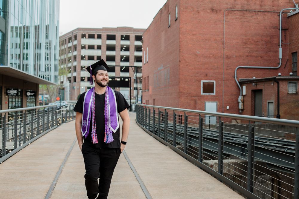 Graduate in academic regalia walking along modern urban walkway with buildings in background.