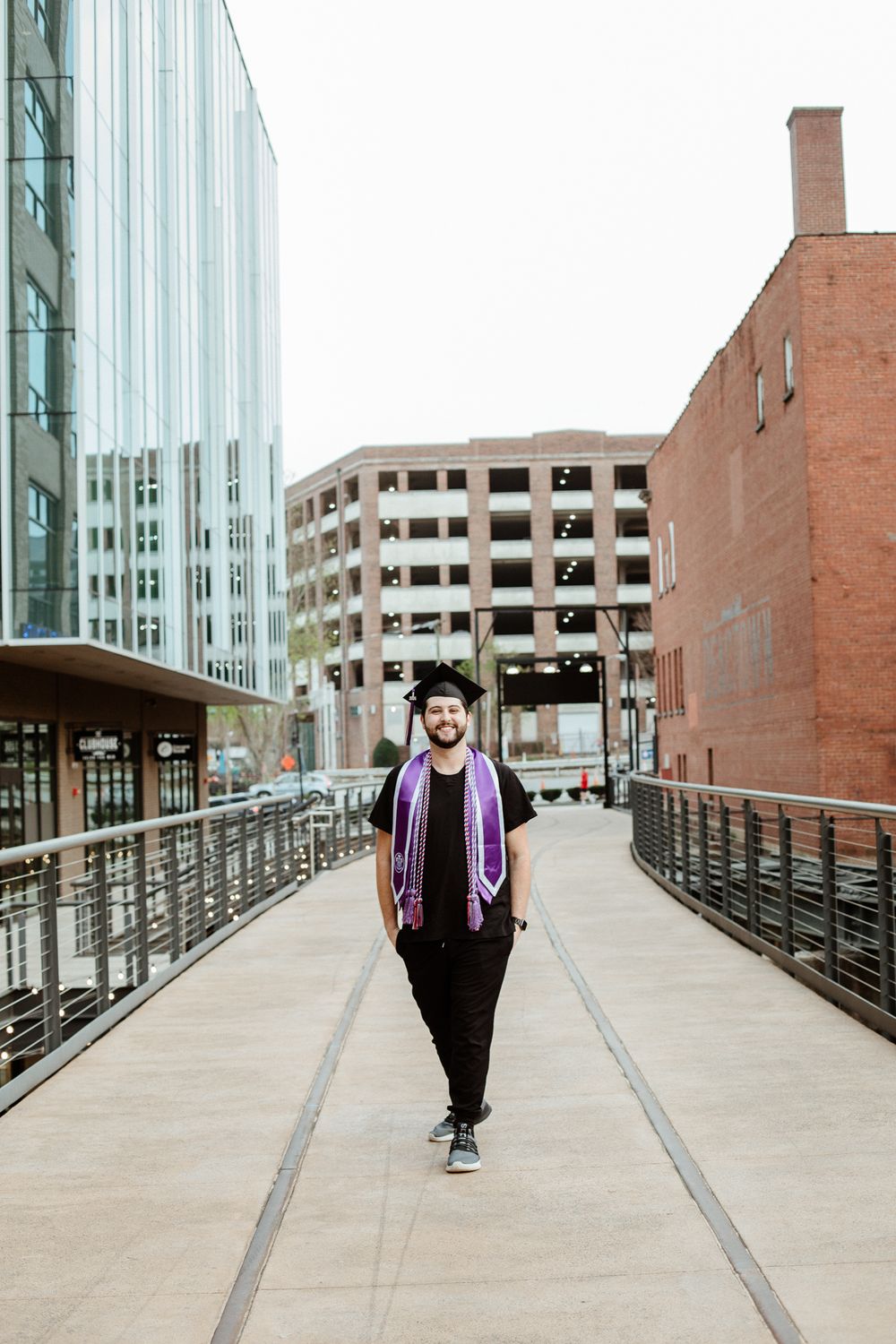 Graduate in academic regalia walking along an elevated walkway with modern buildings in background.