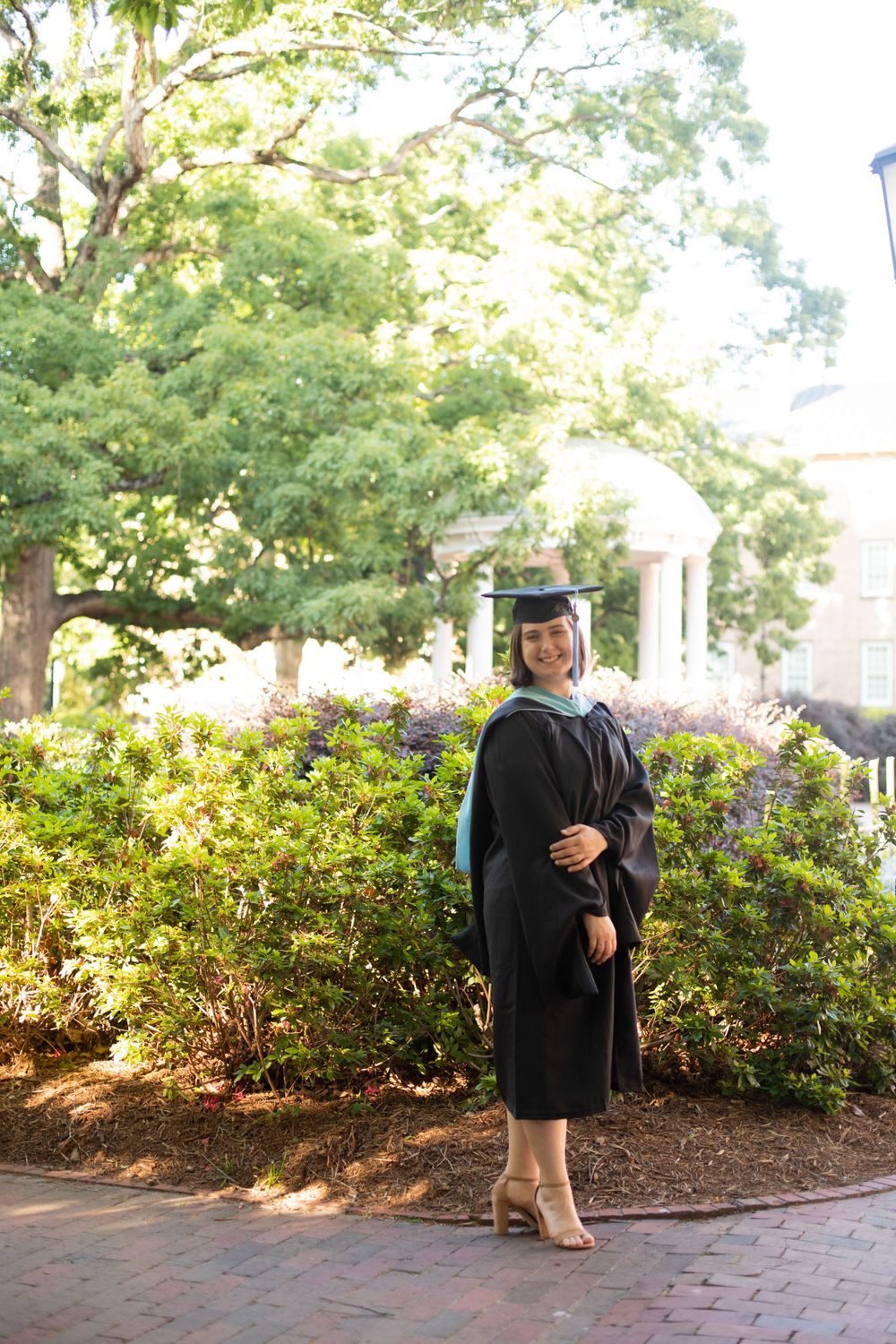 Graduate in black cap and gown poses near flowering bushes on campus path.