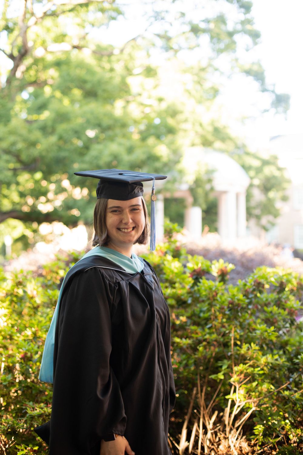 Graduate in traditional black academic regalia poses in garden setting.