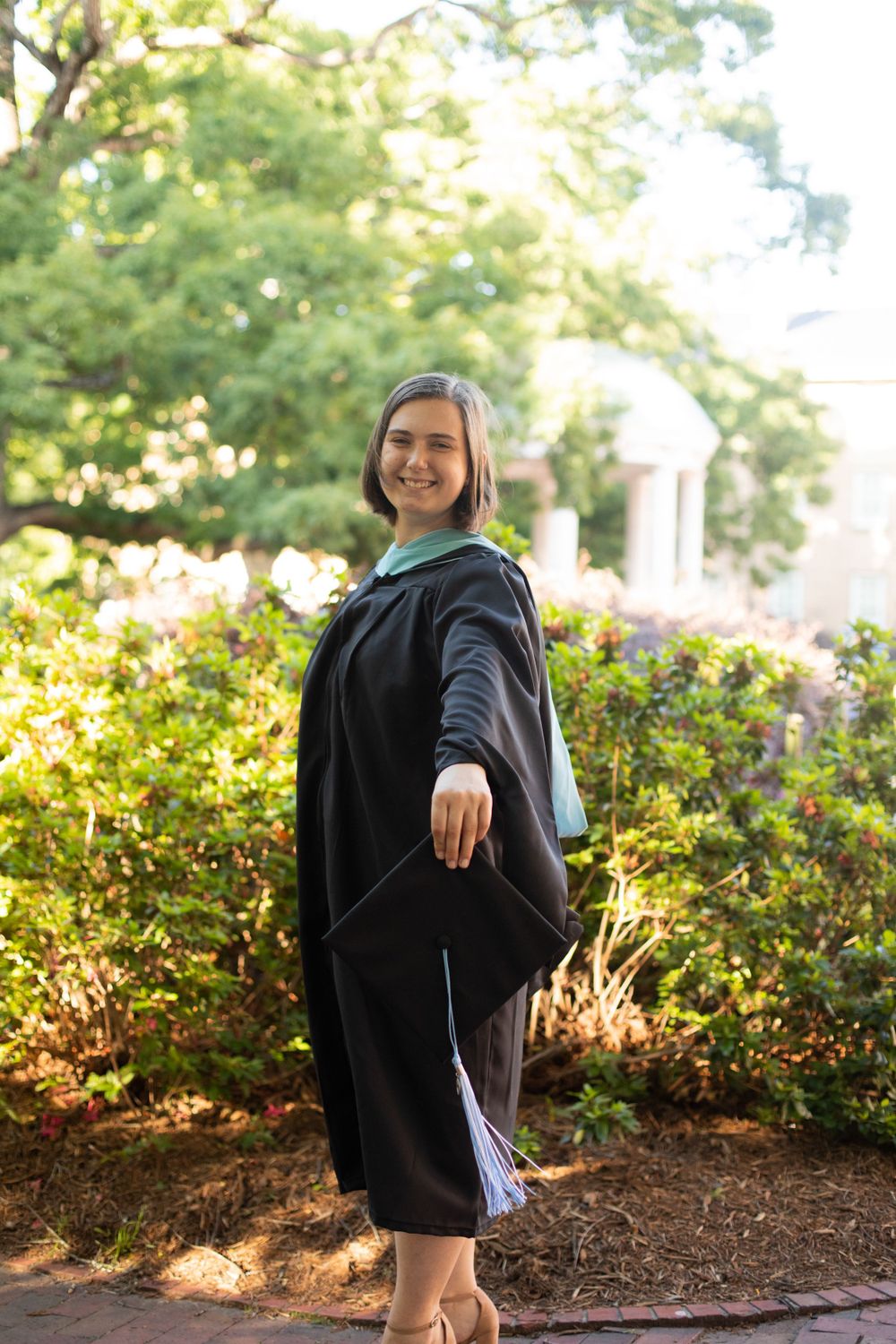 Graduate in black commencement attire poses playfully in garden setting.