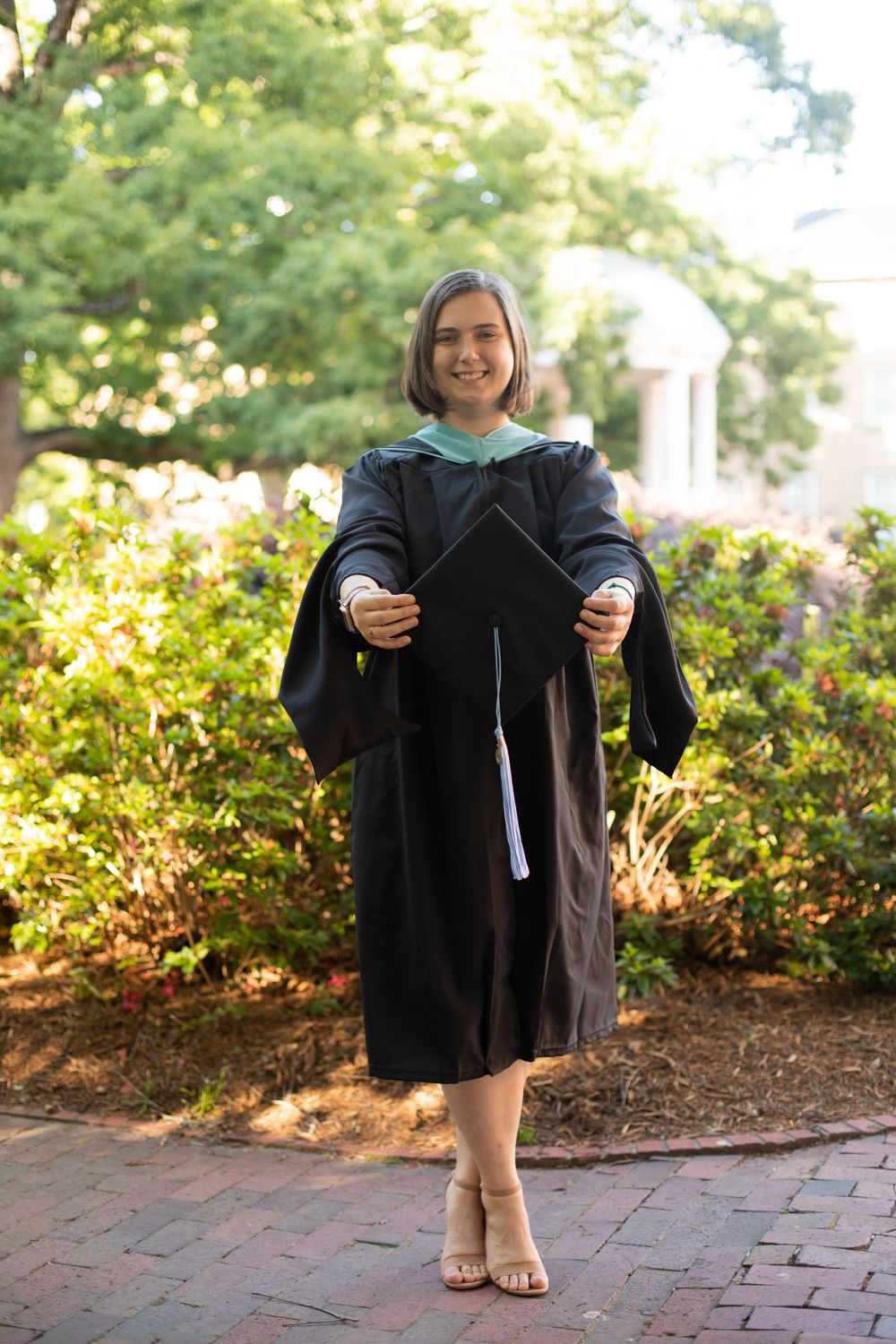 Graduate in black academic regalia poses on brick pathway near garden.