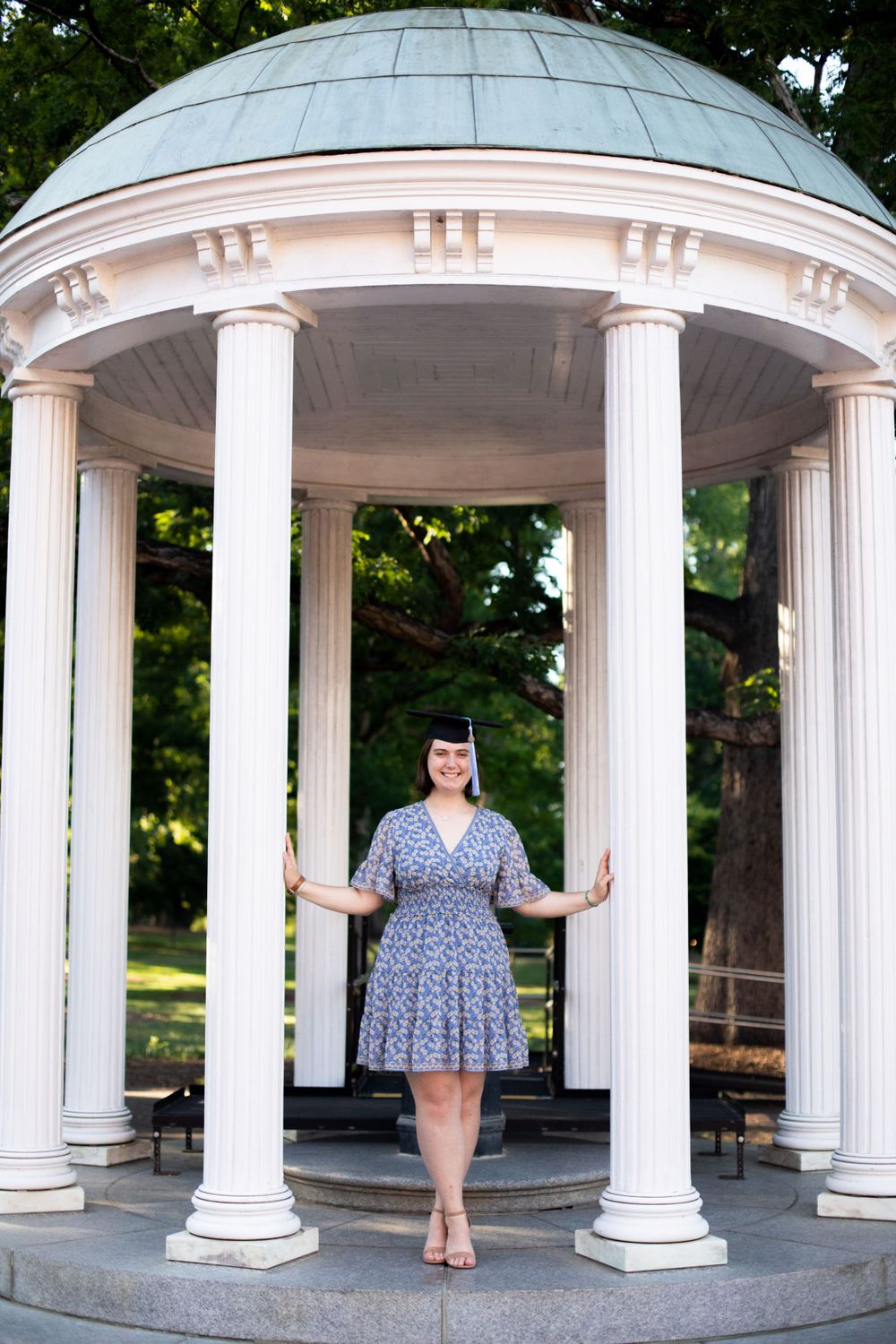 Graduate in blue floral dress poses at UNC Chapel Hill's old well.