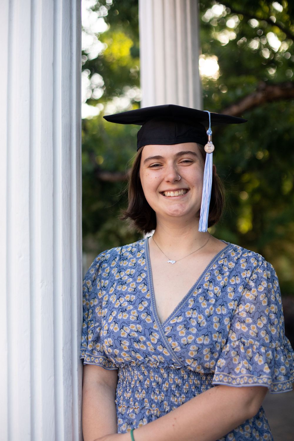 Graduate wearing floral dress and mortarboard poses against white columns.