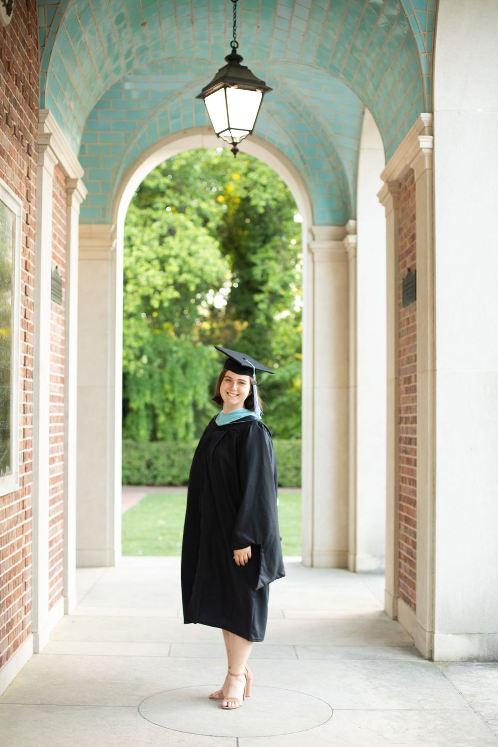 Graduate poses in cap and gown under arched walkway with mint green ceiling.